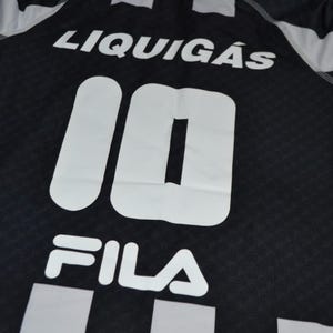 Puede incluir: Camiseta deportiva en blanco y negro con la palabra "LIQUIG&Aacute;S" en la parte superior. El n&uacute;mero "10" se muestra prominentemente en blanco, junto con el logotipo "FILA" en la parte inferior. La camiseta tiene un patr&oacute;n texturizado.