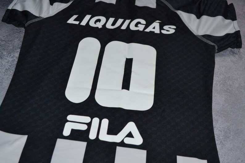 Puede incluir: Camiseta deportiva en blanco y negro con la palabra "LIQUIG&Aacute;S" en la parte superior. El n&uacute;mero "10" se muestra prominentemente en blanco, junto con el logotipo "FILA" en la parte inferior. La camiseta tiene un patr&oacute;n texturizado.