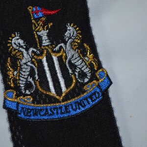 May include: Embroidered Newcastle United crest patch. The crest features a shield with a black and white design, flanked by two seahorses, and a blue banner reading "NEWCASTLE UNITED". The patch is set against a black and white striped background.