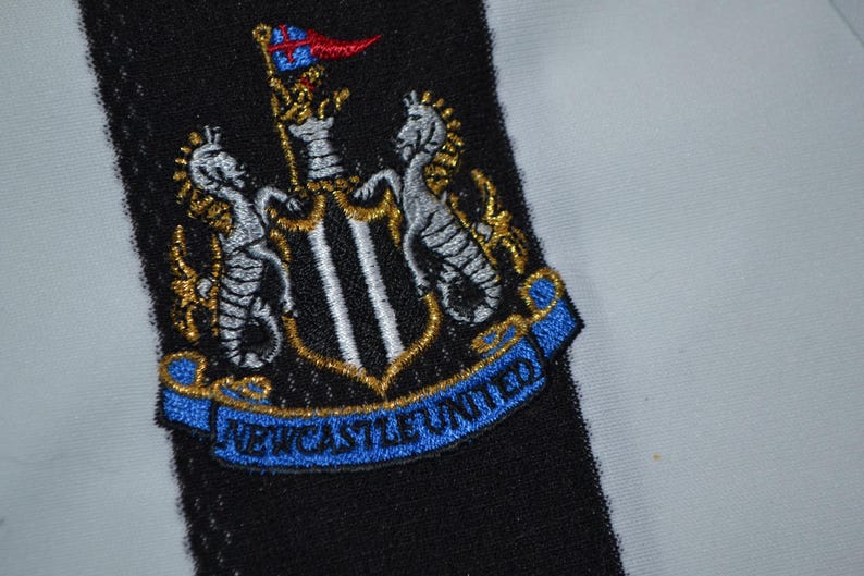 May include: Embroidered Newcastle United crest patch. The crest features a shield with a black and white design, flanked by two seahorses, and a blue banner reading "NEWCASTLE UNITED". The patch is set against a black and white striped background.