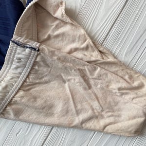 Puede incluir: Primer plano de una prenda beige con cintura blanca y tela azul marino. La tela beige est&aacute; doblada, revelando la cintura blanca con un ribete azul marino. El fondo es una superficie de madera blanca.