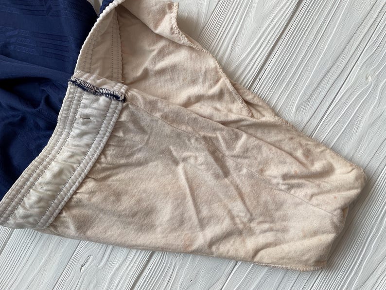 Puede incluir: Primer plano de una prenda beige con cintura blanca y tela azul marino. La tela beige est&aacute; doblada, revelando la cintura blanca con un ribete azul marino. El fondo es una superficie de madera blanca.