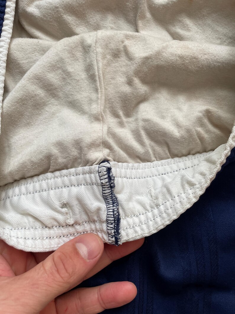 Puede incluir: Primer plano del interior de unos pantalones cortos deportivos azul marino. Los pantalones cortos tienen una cintura el&aacute;stica blanca con cord&oacute;n y un forro interior suave y claro. La tela tiene un aspecto texturizado.