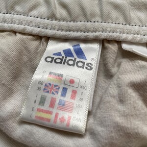Puede incluir: Primer plano de una etiqueta de ropa Adidas. La etiqueta presenta el logotipo de Adidas en azul y blanco, junto con informaci&oacute;n sobre la talla y el pa&iacute;s de origen. El fondo es de tela beige claro.