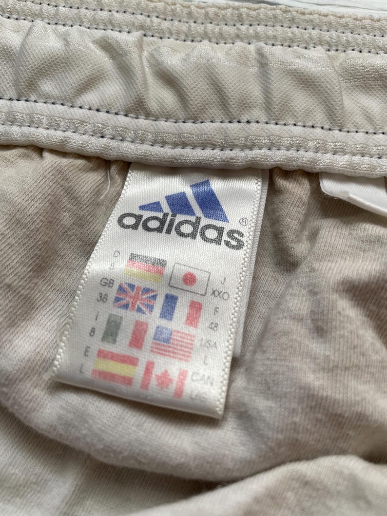 Puede incluir: Primer plano de una etiqueta de ropa Adidas. La etiqueta presenta el logotipo de Adidas en azul y blanco, junto con informaci&oacute;n sobre la talla y el pa&iacute;s de origen. El fondo es de tela beige claro.