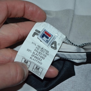 Puede incluir: Primer plano de una etiqueta de ropa con el logotipo de Fila. La etiqueta indica la composici&oacute;n del tejido: 91% poli&eacute;ster y 9% elastano. La talla es M. La prenda es una camiseta deportiva con paneles negros, blancos y grises.