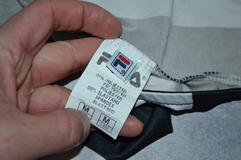 Puede incluir: Primer plano de una etiqueta de ropa con el logotipo de Fila. La etiqueta indica la composici&oacute;n del tejido: 91% poli&eacute;ster y 9% elastano. La talla es M. La prenda es una camiseta deportiva con paneles negros, blancos y grises.