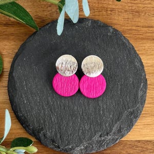 May include: A pair of earrings featuring two stacked circular discs. The top disc is metallic silver with a brushed texture, while the bottom disc is a vibrant hot pink with a fine, vertical ribbed texture. The earrings are circular in shape.
