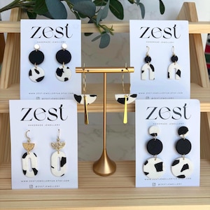 May include: A selection of handmade earrings on display. The earrings showcase black and white patterns, gold details, and diverse shapes, such as circles and arches. The brand name "zest" is printed on the cards.