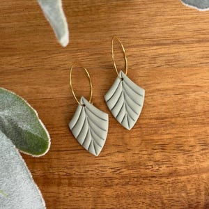 May include: A pair of sage green leaf-shaped earrings with gold-coloured hoop closures. The earrings have a textured, veined design and are displayed on a wooden surface. The earrings are a nature-inspired accessory.