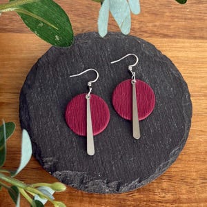 May include: Burgundy-coloured earrings featuring a circular design and silver-coloured vertical bar accents. The earrings are displayed on a dark grey, textured surface, with a wooden background and green foliage.