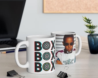 May include: A white ceramic mug featuring the text BOX BOX BOX with the letter O replaced by a Formula 1 tire graphic. The other side of the mug displays a photographic print of a person wearing sunglasses and a racing suit with the text Highland Spring.