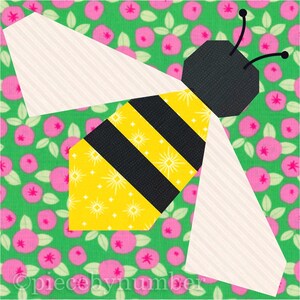 Square quilt block design of a stylized honey bee with embroidered antennae. Bee is yellow and black striped, with cream wings. Background is pink and green floral.