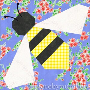 Square quilt block design of a stylized honey bee with embroidered antennae. Bee is yellow and black striped, with white wings. Background is blue and pink floral.