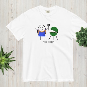 May include: White t-shirt with a cartoon illustration of an egg wearing a blue apron and holding a spatula in front of a green grill. The text "PRO CHEF" is below the illustration.