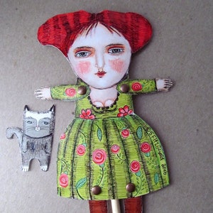 May include: A paper doll with red hair, a green dress with red roses, and brown boots. The doll is holding a stick and has a small gray cat beside it. The doll's dress has the text "S. FitzGerald 2016" on it.
