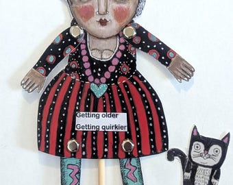 May include: A paper puppet of a person with a gray patterned head covering, a black dress with red vertical stripes, and teal boots with pink lightning bolts. The dress features the text Getting older Getting quirkier and a small black cat.