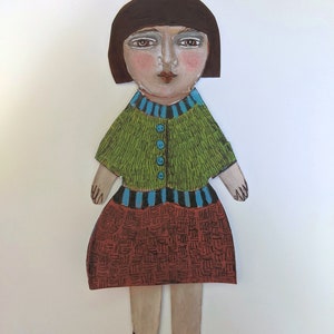 May include: A paper doll with brown hair, a green sweater with blue buttons, a striped waistband, and a brown and black patterned skirt. The doll is wearing black shoes.