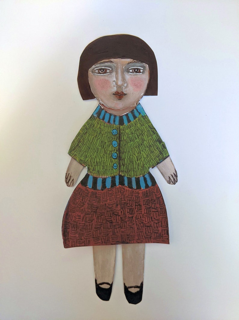 May include: A paper doll with brown hair, a green sweater with blue buttons, a striped waistband, and a brown and black patterned skirt. The doll is wearing black shoes.