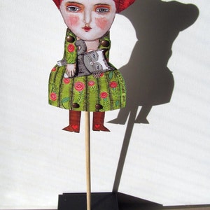 May include: A handmade wooden puppet of a woman with red hair wearing a green dress with red flowers and holding a gray cat. The puppet is on a wooden stick with a black base. The base has the text "OSF Handmade 2016" on it.