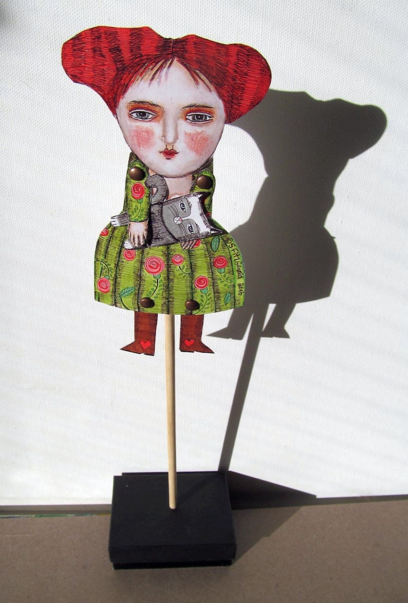 May include: A handmade wooden puppet of a woman with red hair wearing a green dress with red flowers and holding a gray cat. The puppet is on a wooden stick with a black base. The base has the text "OSF Handmade 2016" on it.
