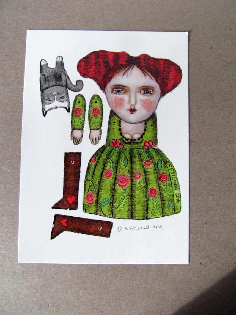 May include: A paper doll with red hair, a green dress with red roses, and brown shoes. The doll is holding a cat in its arms. The doll is part of a paper doll set. The image includes the text "S.Fitzgerald 2018".