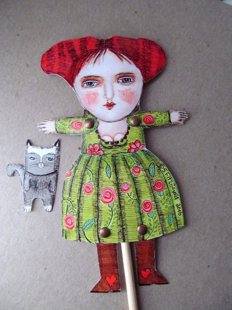 May include: A paper doll with red hair, a green dress with red roses, and brown boots. The doll is holding a stick and has a small gray cat beside it. The doll's dress has the text "S. FitzGerald 2016" on it.