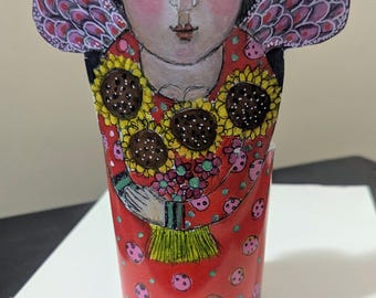 May include: A cylindrical decorative figure featuring a painted illustration of a person with black hair, a floral crown, and wings. The figure wears a red garment adorned with pink polka dots and holds a bouquet of yellow sunflowers.