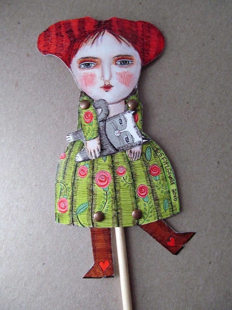 May include: A hand-painted paper doll with red hair, a green dress with red roses, and brown boots. The doll is holding a gray cat and is attached to a wooden stick. The doll is signed "StiffGweed 2016".