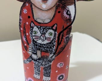 May include: A cylindrical paper craft figure featuring a person with brown hair wearing a red dress with pink floral patterns, holding a black and white cat. The figure holds a yellow balloon with the word Hope written in black ink.