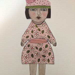 May include: A paper doll with a pink dress with white flowers and green leaves, a pink and white polka dot hat with a pink flower, and black shoes.
