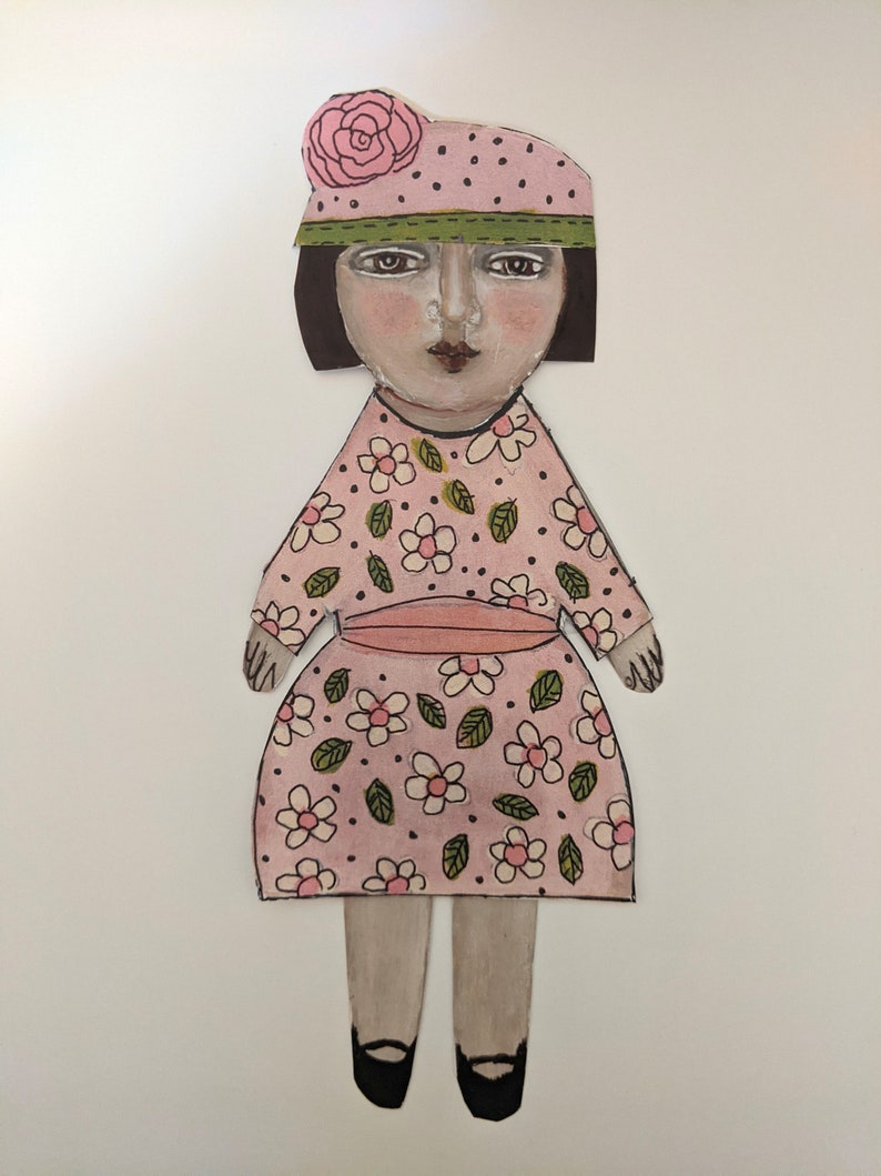 May include: A paper doll with a pink dress with white flowers and green leaves, a pink and white polka dot hat with a pink flower, and black shoes.