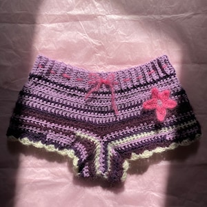 May include: Purple and white crocheted shorts with a pink flower detail. The shorts have a ruffled hem and a tie at the waist.