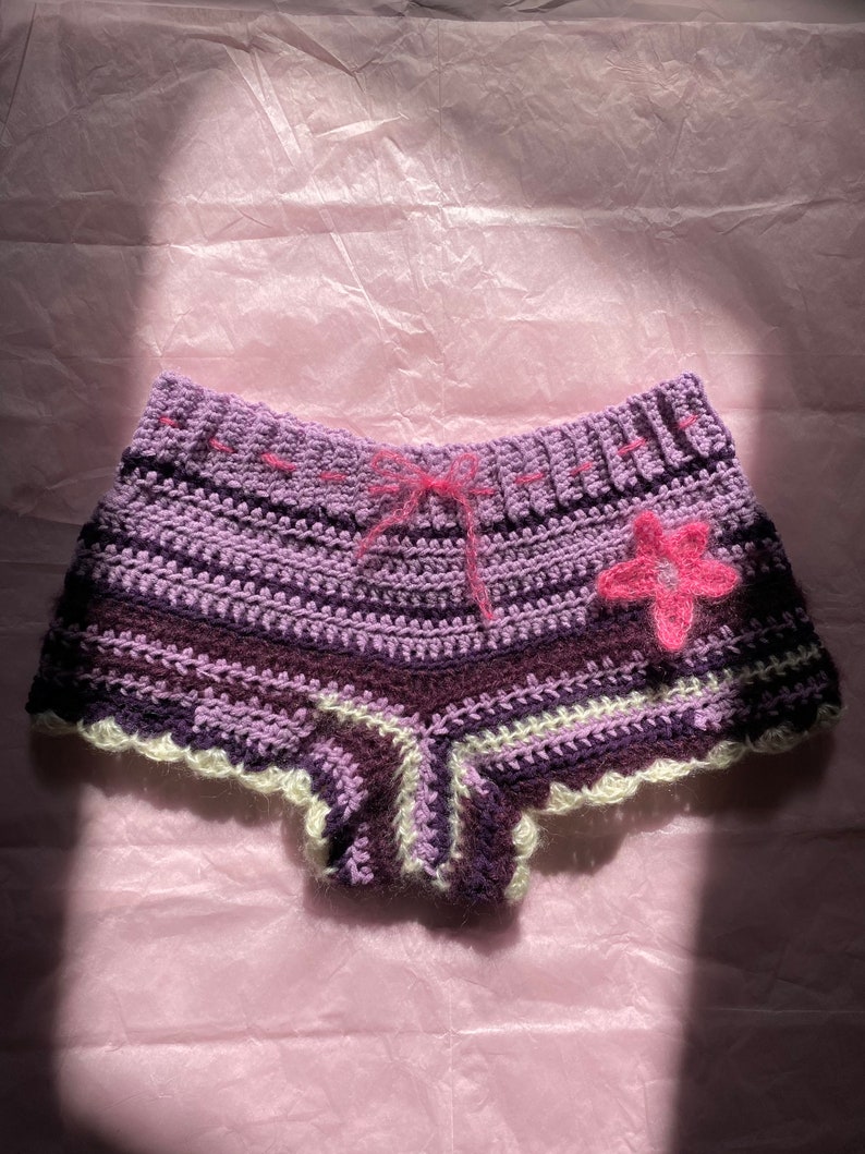 May include: Purple and white crocheted shorts with a pink flower detail. The shorts have a ruffled hem and a tie at the waist.