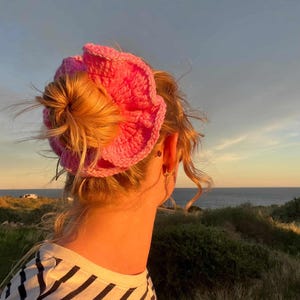 May include: A woman with blonde hair wearing a pink crocheted headband. The headband is in the shape of a flower. The woman is wearing a black and white striped shirt. The text "@lissis.crochet" is in the top left corner.
