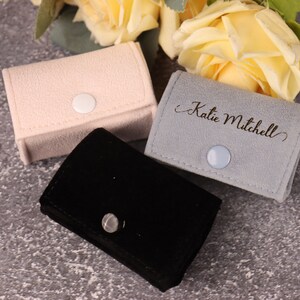 May include: Three small, rectangular velvet boxes with snap closures. The boxes are in pink, gray, and black. The gray box has the name "Katie Mitchell" engraved on it.