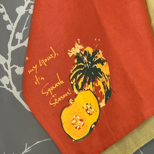 May include: A red kitchen towel with a pumpkin illustration and the text "Oh my gourd, it's Squash Season!". The pumpkin is yellow and green. The towel is folded, with a gray and white floral pattern in the background.
