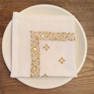 May include: White linen napkin with a gold floral design and small sequins, on a white plate. The napkin has a decorative border and floral accents, suitable for elegant dining.