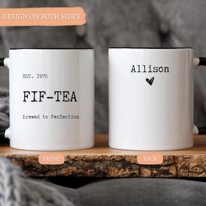 May include: Two white ceramic mugs with black handles and rims. One mug reads "EST. 1975 FIF-TEA Brewed to Perfection." The other mug has the name "Allison" and a heart symbol. The mugs are on a wooden surface.
