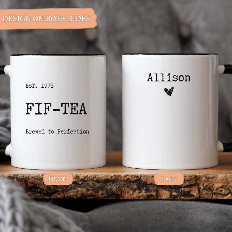 May include: Two white ceramic mugs with black handles and rims. One mug reads "EST. 1975 FIF-TEA Brewed to Perfection." The other mug has the name "Allison" and a heart symbol. The mugs are on a wooden surface.