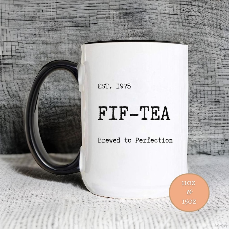 May include: White ceramic mug with a black handle and rim. The mug features the text "EST. 1975 FIF-TEA Brewed to Perfection". A small orange circle indicates the mug is available in 11oz and 15oz sizes.