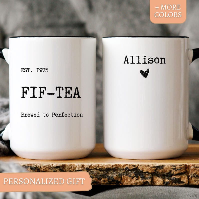 May include: Two white ceramic mugs with black rims and handles. One mug reads "EST. 1975 FIF-TEA Brewed to Perfection." The other mug says "Allison" with a heart symbol. The mugs are on a wooden surface. The words "PERSONALIZED GIFT" are at the bottom.