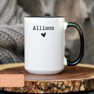 May include: White ceramic mug with a black interior and handle. The mug features the name "Allison" in black text with a small black heart below it. The mug is on a wooden surface.