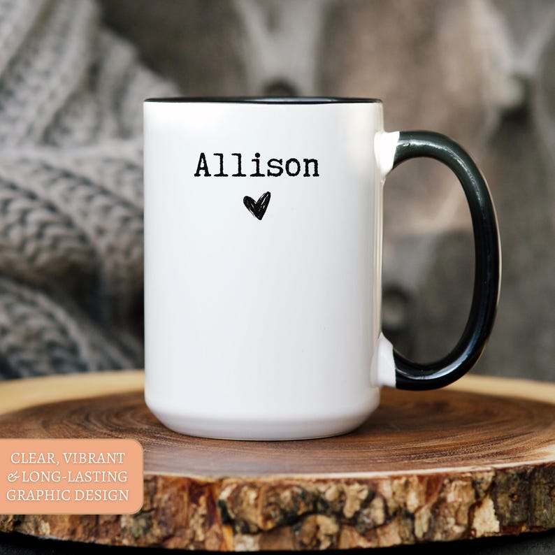 May include: White ceramic mug with a black interior and handle. The mug features the name "Allison" in black text with a small black heart below it. The mug is on a wooden surface.