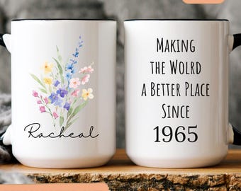 May include: White ceramic mug with a black rim and handle. One side features a watercolour floral design with the name "Racheal". The other side reads "MAKING THE WOLRD A BETTER PLACE SINCE 1965".