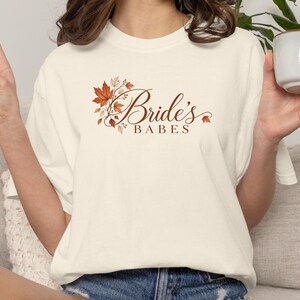 May include: A cream-colored short-sleeve t-shirt featuring a graphic print of autumn maple leaves in shades of orange, brown, and tan. The text "Bride's BABES" is printed in a brown script and serif font centered below the foliage design.