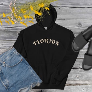 May include: Black hoodie with a cream coloured "FLORIDA" graphic on the front. The hoodie is laid out on a wooden surface with a pair of black sliders and denim shorts.