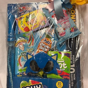 Pode incluir: Uma cesta de presente embrulhada em celofane transparente, amarrada com um laço azul, rosa e amarelo. A cesta contém vários doces, uma varinha de bolhas azul e uma máscara do Sonic. Inclui Jolly Rancher Freezer Pops.