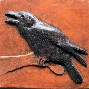 May include: A square terracotta-colored tile featuring a raised, dark brown relief sculpture of a raven perched on a branch. The bird is depicted in profile with its beak slightly open, showcasing a textured surface and detailed wing feathers.