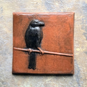 May include: A square terracotta-colored tile featuring a raised, dark black relief of a bird perched on a horizontal line. The surface of the tile has a matte, earthy finish with subtle variations in the reddish-brown tone across the square base.